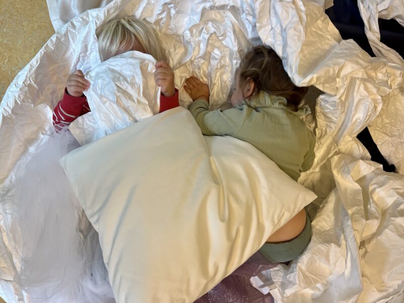 Visual description: Colored photograph from above of two children, respectively 2-3 years old, lying in a pile of light, wrinkled textiles. The children lie next to each other and on top of them lies a large bright pillow. The child on the left is wearing a red sweater, only the sleeves are visible. The child lies on their back and holds a piece of the white fabric over his face. The child on the right is wearing a green jumper and green trousers. The child lies on their stomach with their head down towards the floor, and their body is barely visible from under the large pillow. They both seem to be in motion, in play.