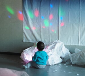 Visual description: Color photograph of a small child sitting on the floor with their back turned. The face is turned towards a light wall where a large white textile hangs, like a curtain. Rays of light in the colors blue, green and pink appear on the textile and the wall next to it. In the area around the child are large, wrinkled formations of white textile or paper. At the very front of the picture we can see some light pink tulle sticking up and into the picture.